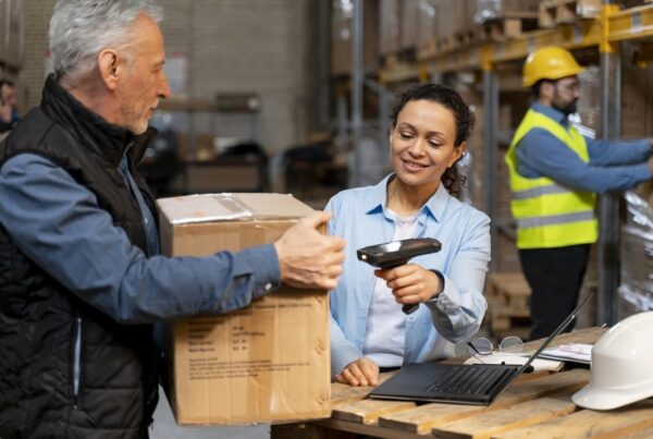 Funcionários em um armazém realizando o processo de entrega e escaneamento de pacotes com um leitor de código de barras, enfatizando a logística e o trabalho em equipe.