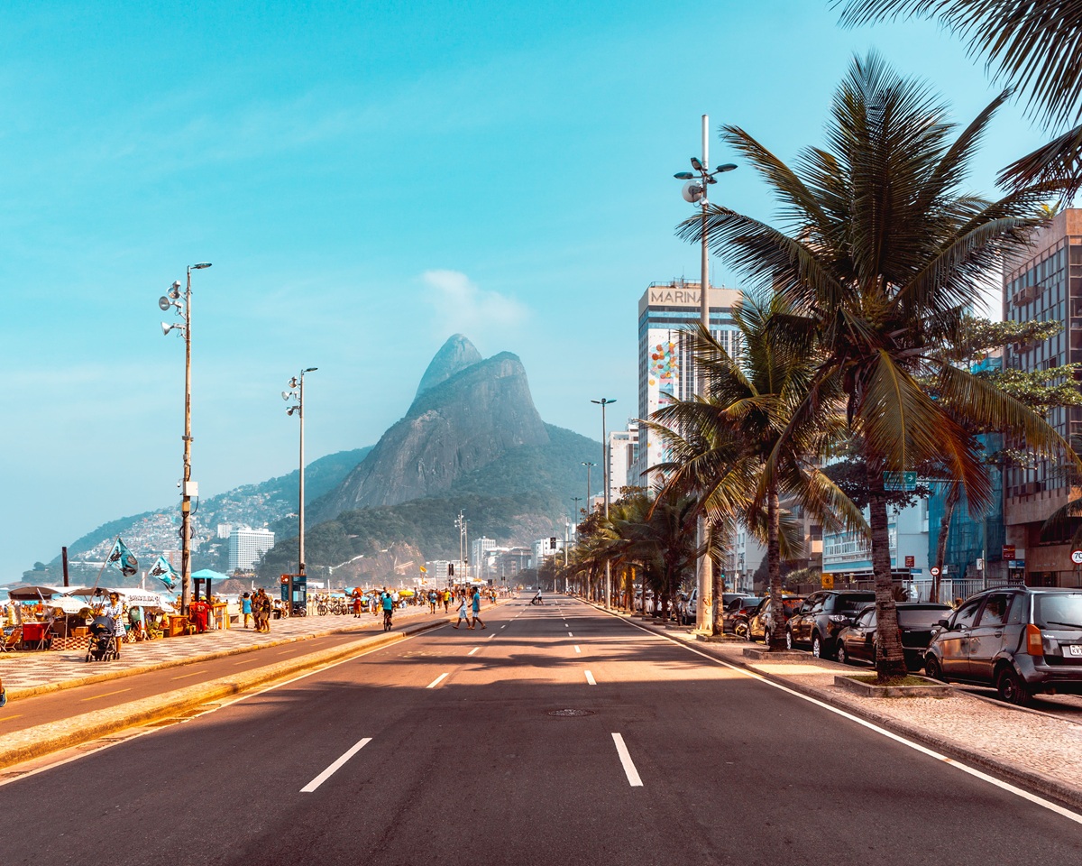 Vista panorâmica da praia de Copacabana no Rio de Janeiro, com palmeiras e o famoso Morro do Pão de Açúcar ao fundo, em um dia ensolarado.
