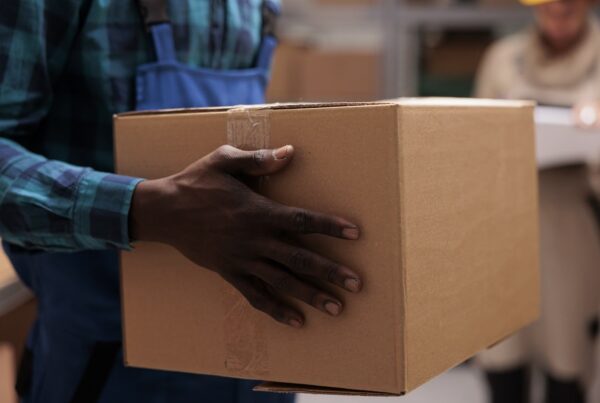 Homem segurando uma caixa de papelão, representando logística e transporte de produtos. A imagem ilustra o trabalho nas entregas e movimentação de mercadorias.
